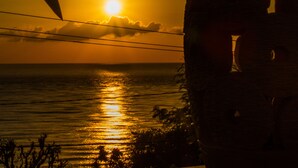 A sunset over the ocean with silhouettes of palm trees and a large rock formation in the foreground. - Beten Waru (Karangasem)