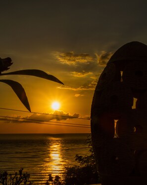 A sunset over the ocean with silhouettes of palm trees and a large rock formation in the foreground. - Beten Waru (Karangasem)