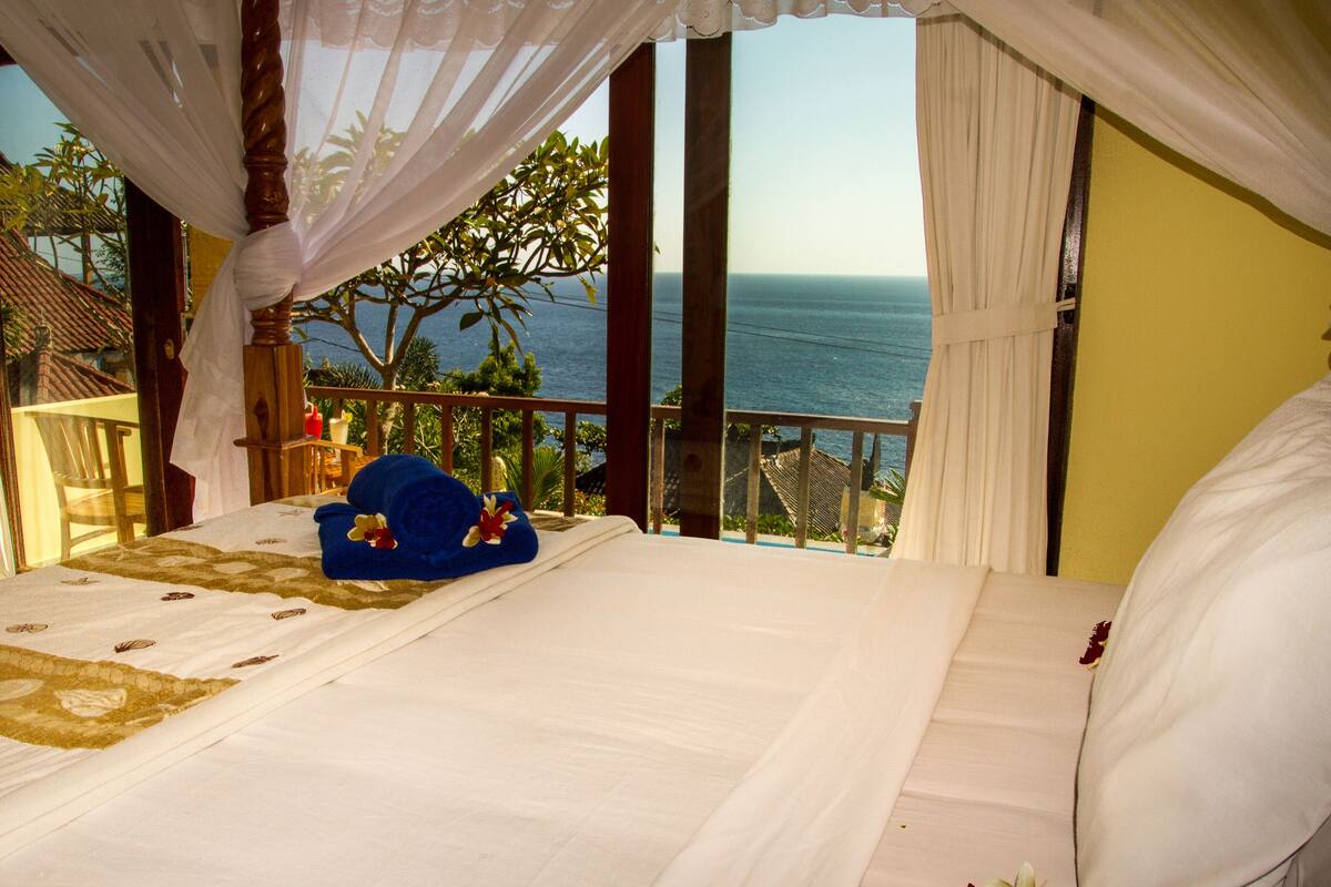 A bedroom with a bed, a view of the ocean, and a balcony with plants.