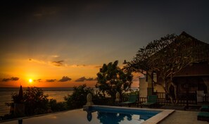 Una piscina con vista al atardecer, un edificio con balcón y un árbol en primer plano.