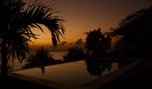 Una puesta de sol serena sobre una piscina tranquila, con palmeras y una vista distante del mar.