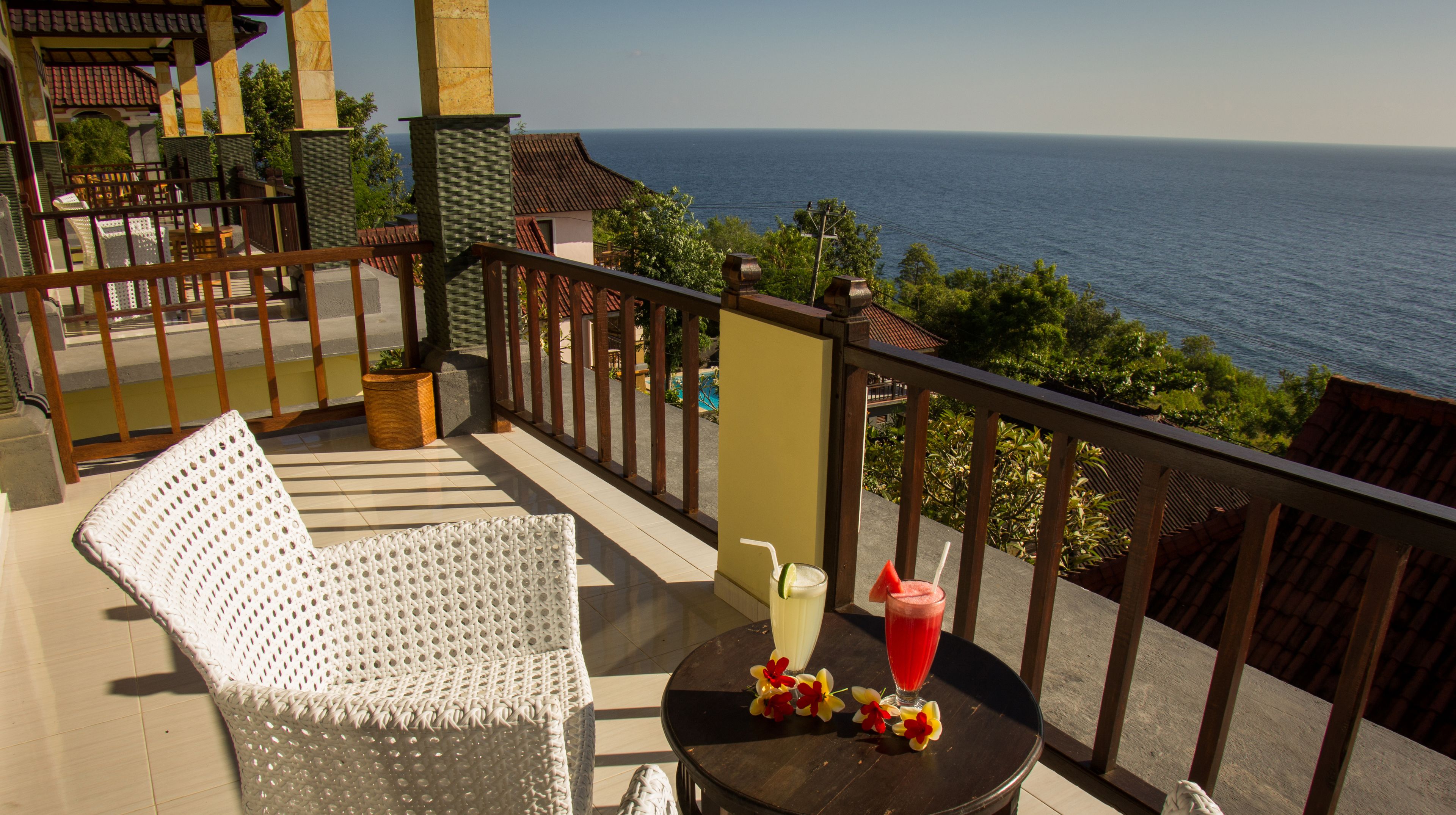 A balcony with a white woven chair, a round table with drinks, and a view of the sea.