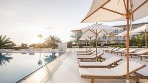 Zona junto a la piscina con sillones de descanso y sombrillas, cielo despejado y un edificio al fondo.