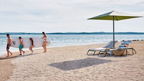 Escena de playa con una fila de personas llevando tablas de surf, una sombrilla y sillas de playa con una bebida en la mesa.