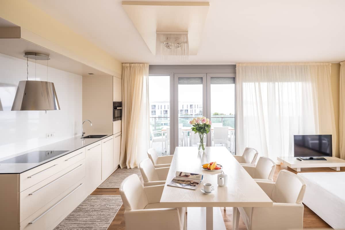 A modern hotel room with a kitchen, dining area, and a large window with curtains.