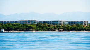 Un hotel moderno con vistas al agua, que presenta un edificio grande con varios pisos y techo plano, rodeado de abundante vegetación y un cielo azul despejado.
