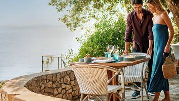 A couple dining at an outdoor table with wicker chairs, a stone wall, and a view of the sea.