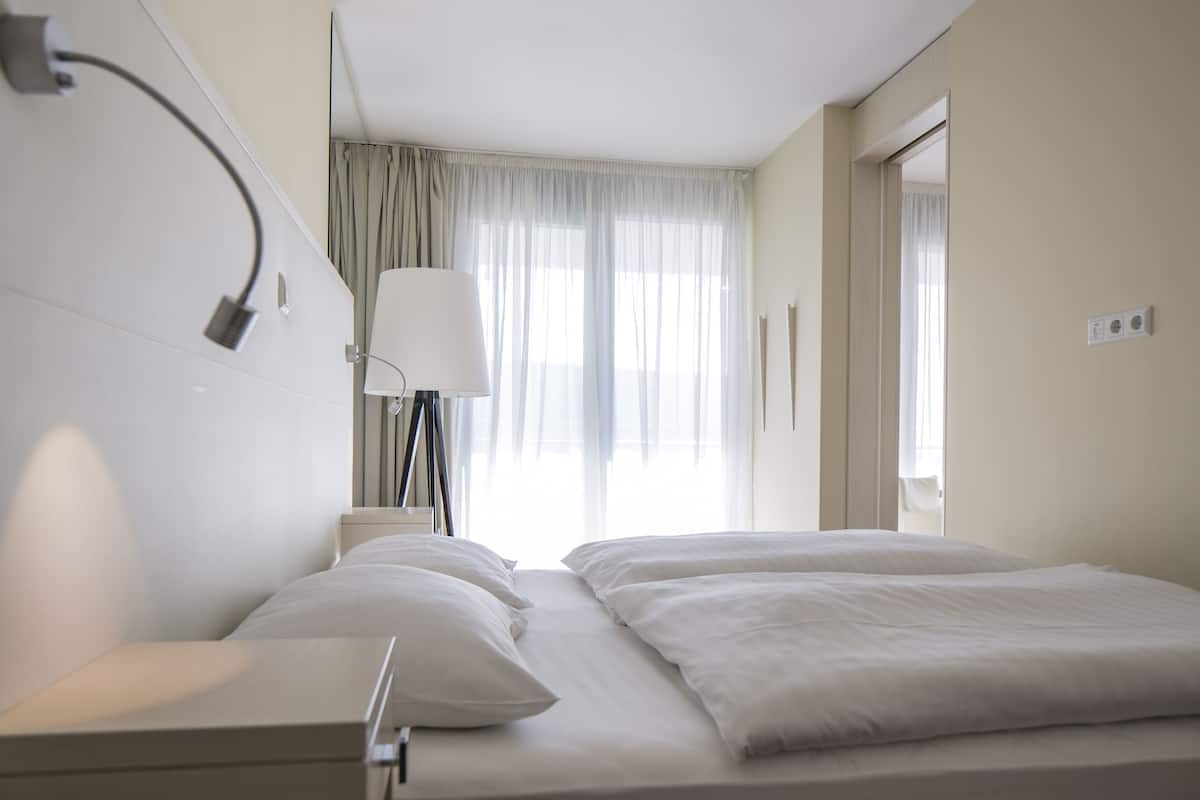 A hotel room with a bed, a bedside lamp, a window with curtains, and a door.