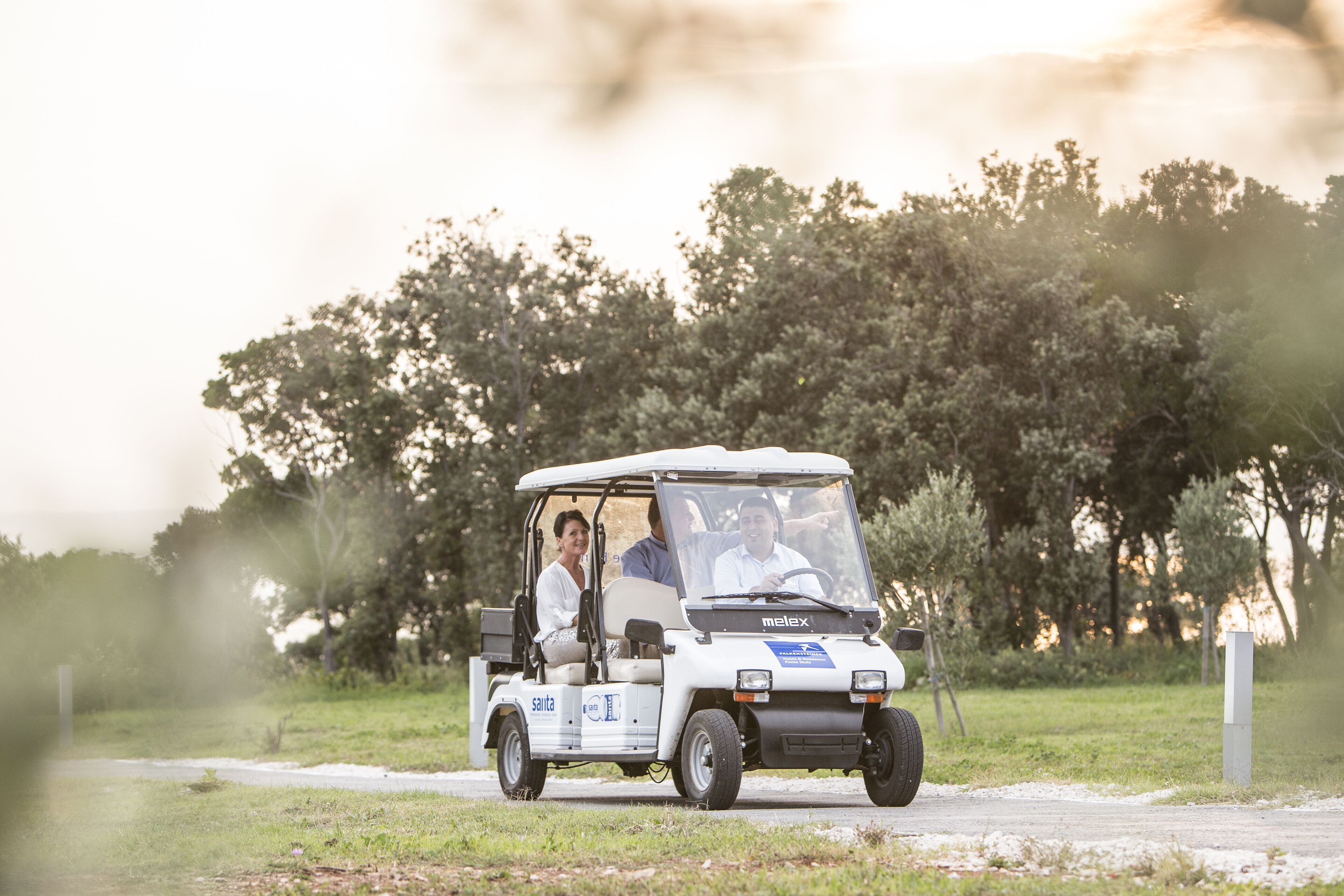 Ein Golfwagen mit drei Personen auf einem befestigten Weg, umgeben von Grünflächen.