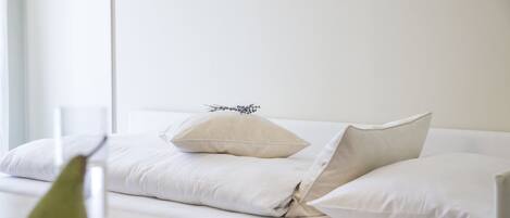 Una cama bien tendida con sábanas y almohadas blancas, una mesita de noche con un objeto verde y una ventana con luz natural.