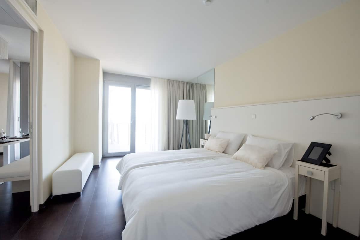 A hotel room with a large bed, a bedside table with a lamp, a bench, and a large window with curtains.