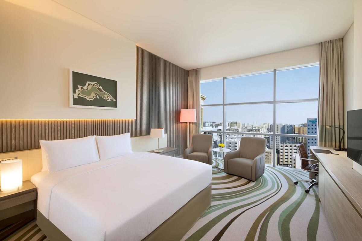 A hotel room with a large bed, a desk with a TV, a chair, and a view of the city through large windows.