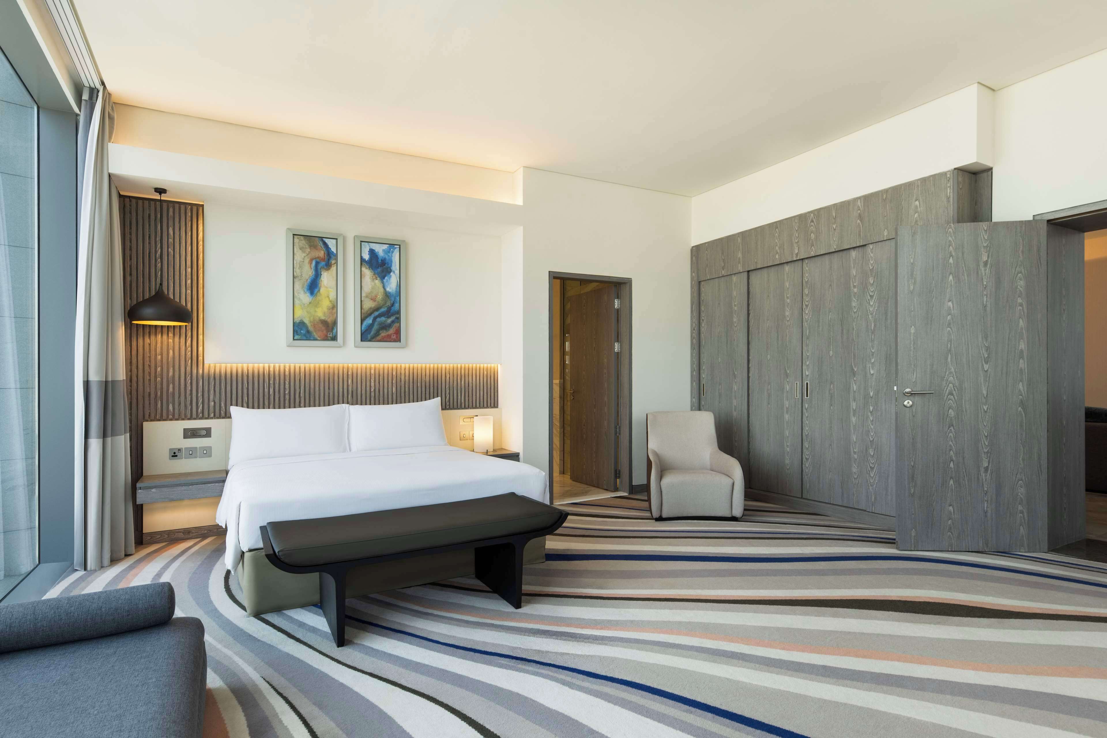 A modern hotel room with a large bed, a bench, a chair, and a wardrobe.