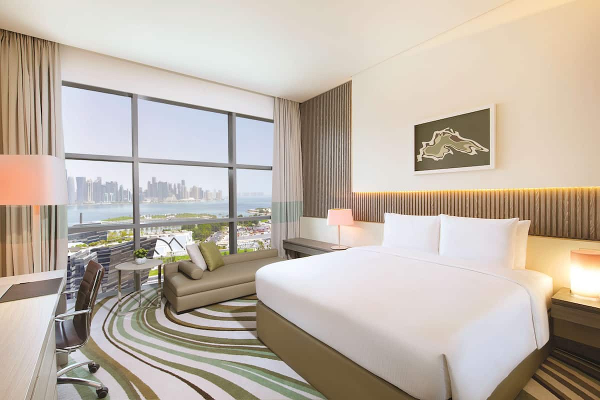 A hotel room with a large bed, a sofa, a desk, and a view of a cityscape through large windows.