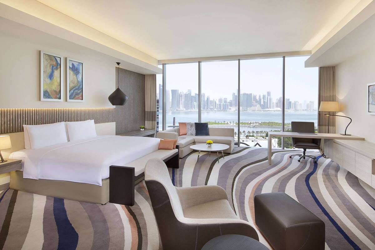 A modern hotel room with a large bed, a sofa, a desk, and a view of a city skyline.