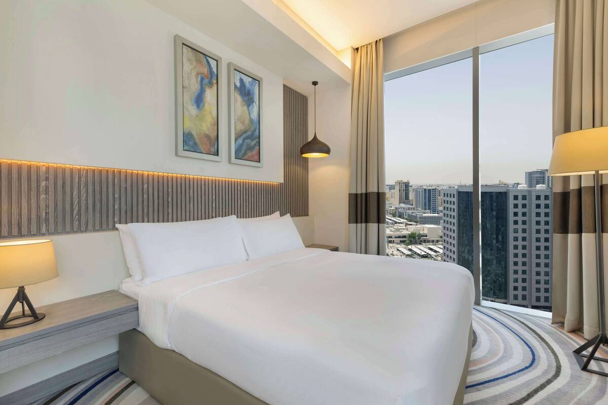 A hotel room with a large bed, a nightstand with a lamp, a wall-mounted artwork, and a view of the city through large windows.