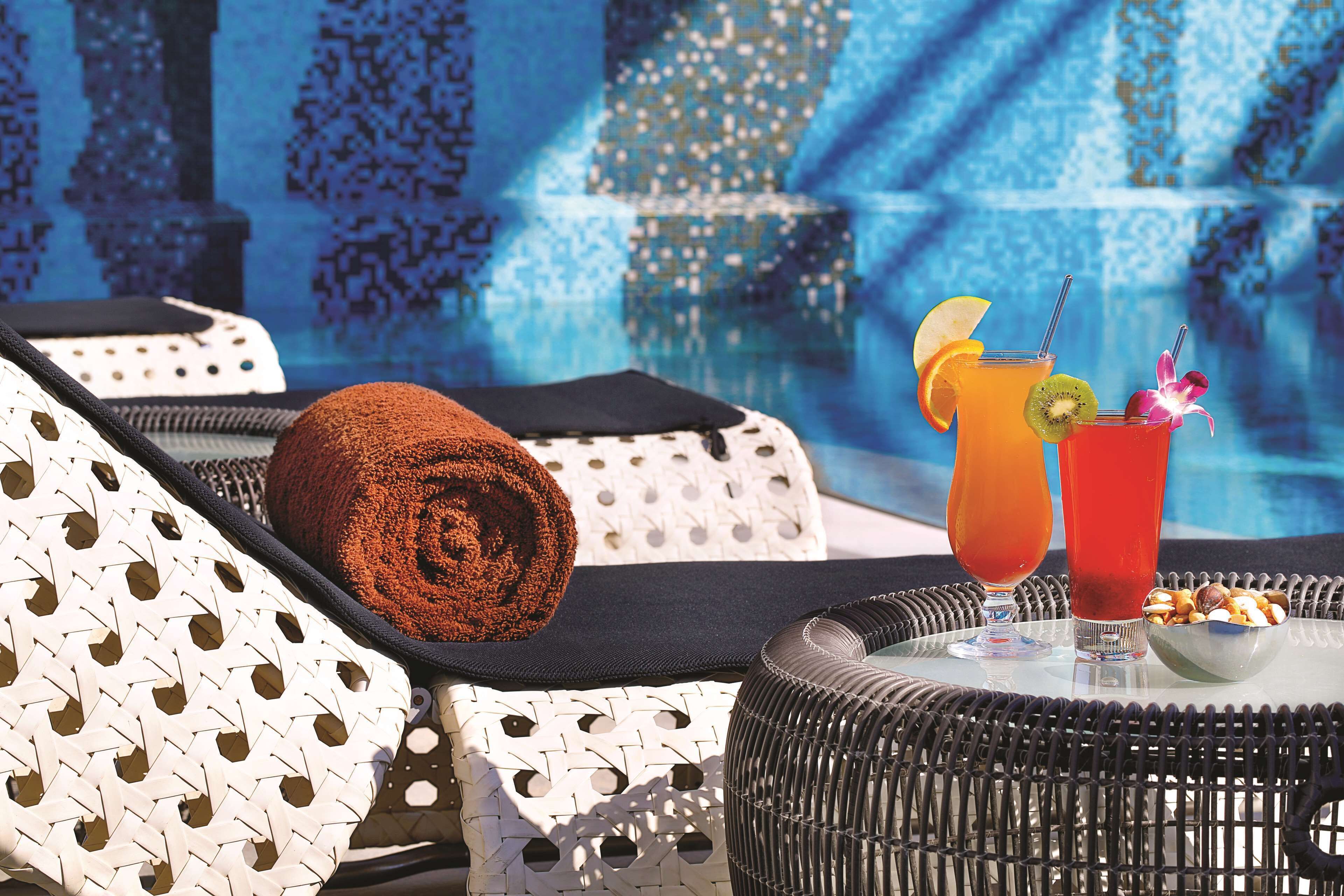 A poolside lounge area with wicker furniture, a towel, two drinks with garnishes, and a bowl of nuts.
