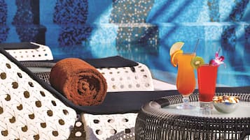 A poolside lounge area with wicker furniture, a towel, two drinks with garnishes, and a bowl of nuts.