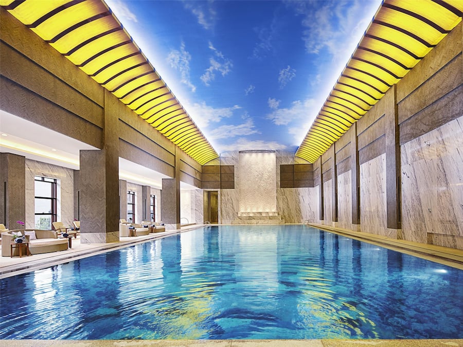 An indoor swimming pool with a ceiling that mimics a sky and clouds, surrounded by walls with herringbone patterns and marble.