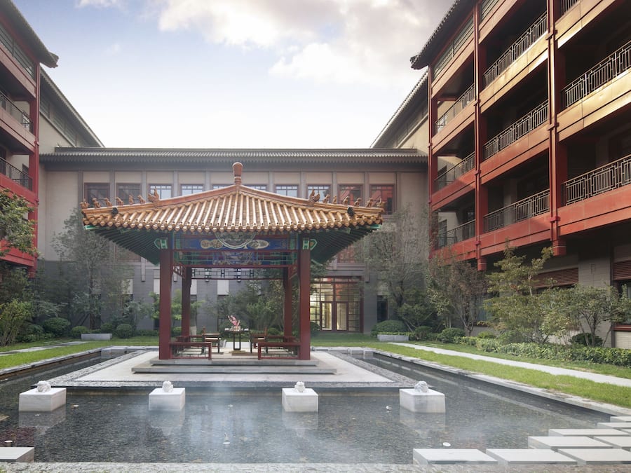 A traditional pavilion with a tiled roof, surrounded by a water feature and stone slabs, set against a backdrop of modern buildings with balconies.