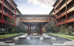 Un pavillon traditionnel, doté d’un toit en tuiles, entouré d’un bassin et de dalles de pierre, se dressant sur un fond de bâtiments modernes dotés de balcons.