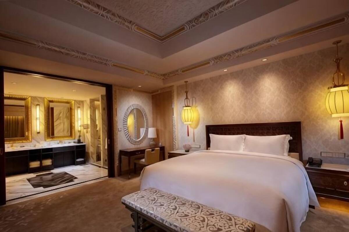 Une chambre spacieuse avec un grand lit, une banquette, une coiffeuse avec un miroir et deux lampes fixées au mur.