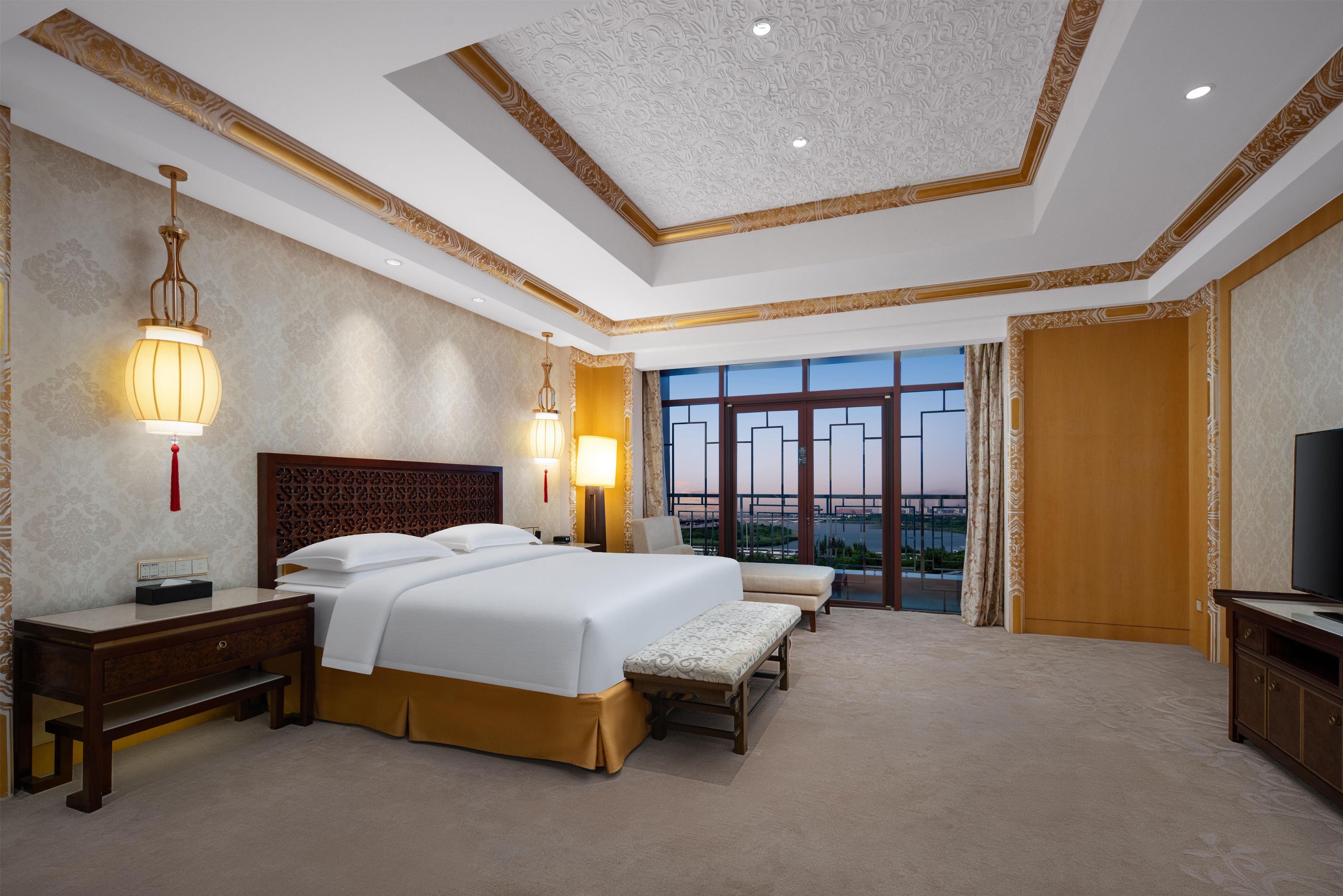 Habitación de hotel con una cama grande, mesitas de noche, televisor y un balcón con vista a la ciudad.