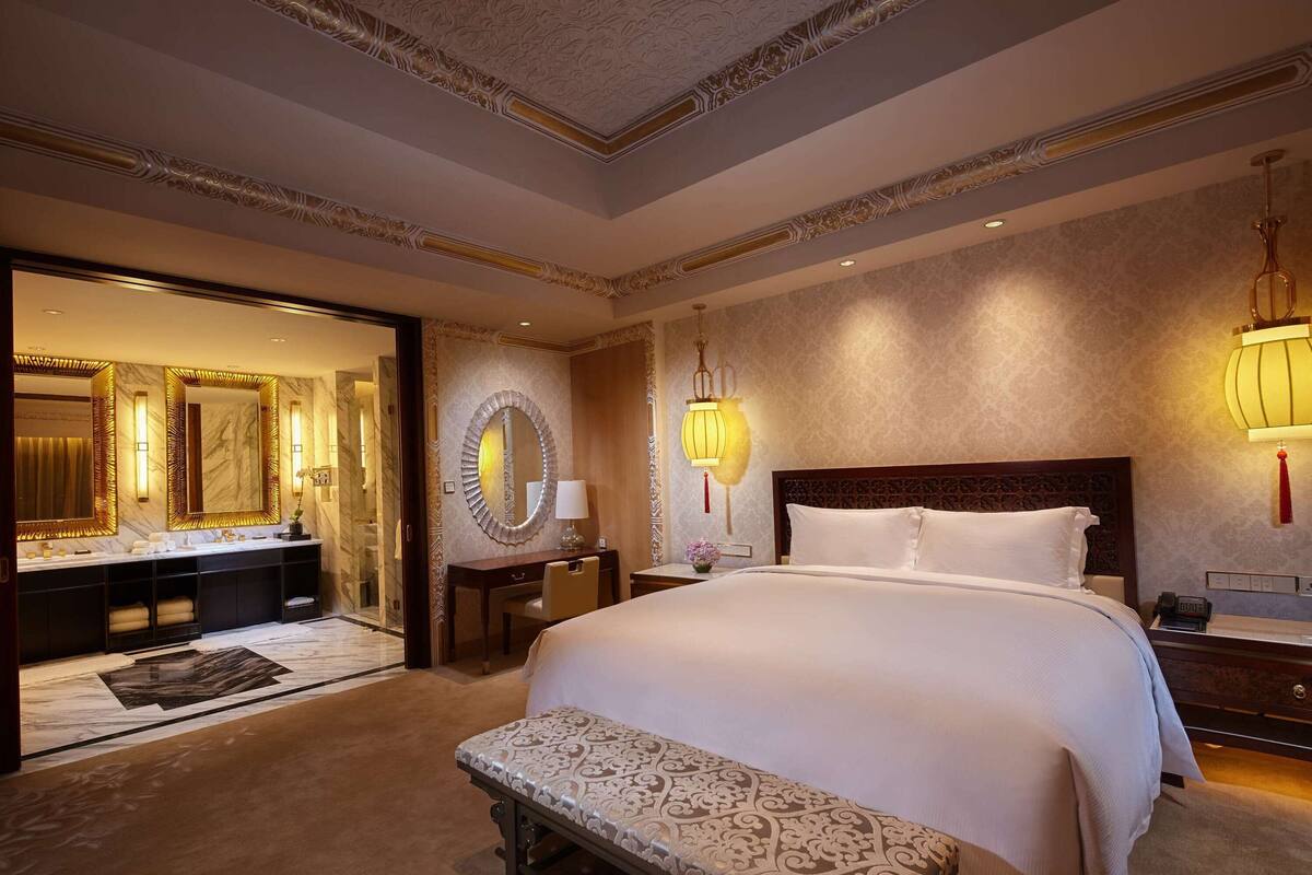 Une chambre d’hôtel luxueuse, dotée d’un grand lit, d’une banquette, d’une coiffeuse avec un miroir et d’une salle de bain bien éclairée.