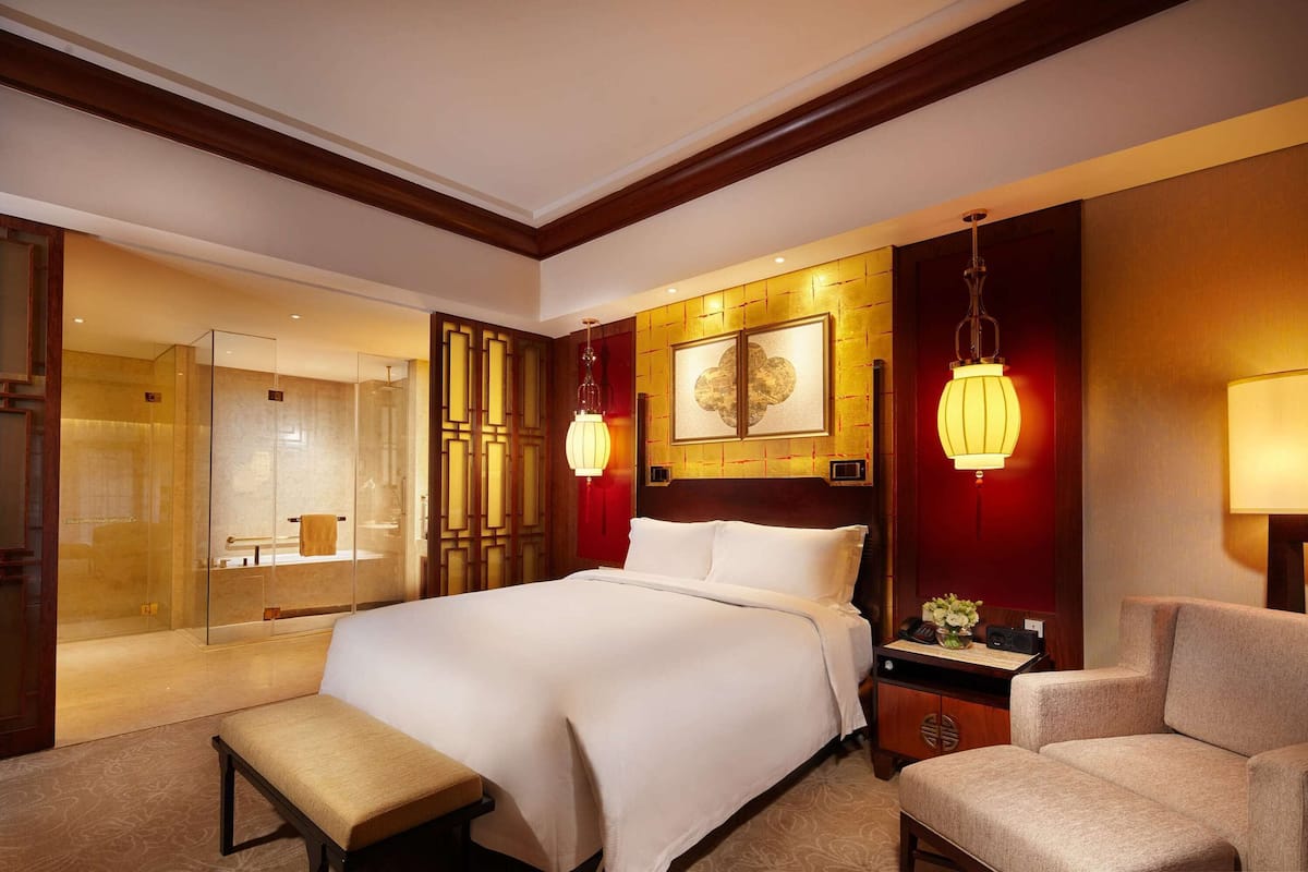 Une chambre d’hôtel avec un grand lit, une table de chevet avec une lampe, une chaise et une cabine de douche en verre.