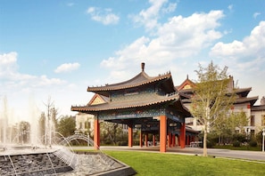 Un pavillon chinois traditionnel, doté d’un toit en tuiles, entouré d’une fontaine et de verdure.