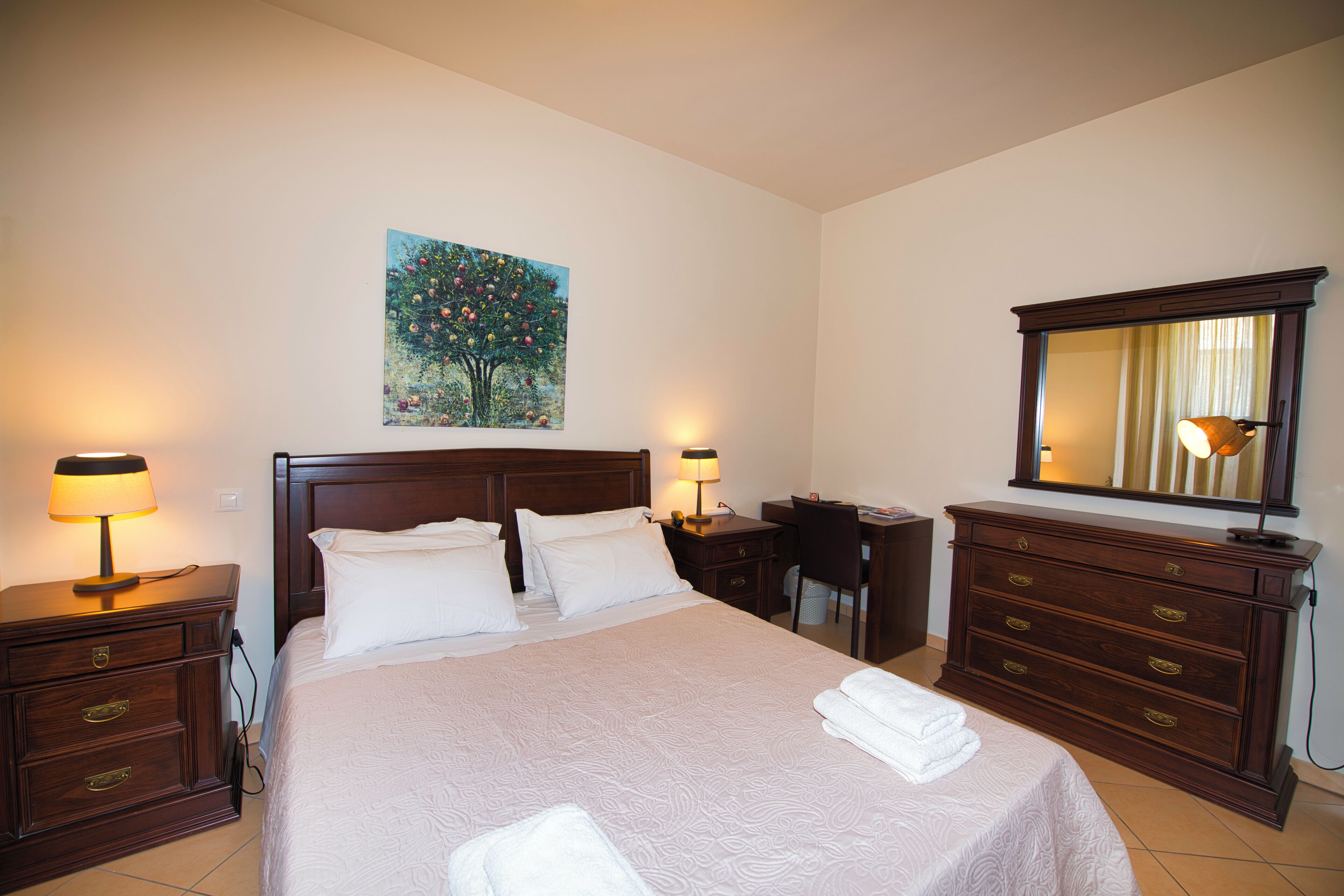 Quarto de hotel com uma cama grande, duas mesas de cabeceira com luminárias, uma escrivaninha, um aparador e um quadro na parede.