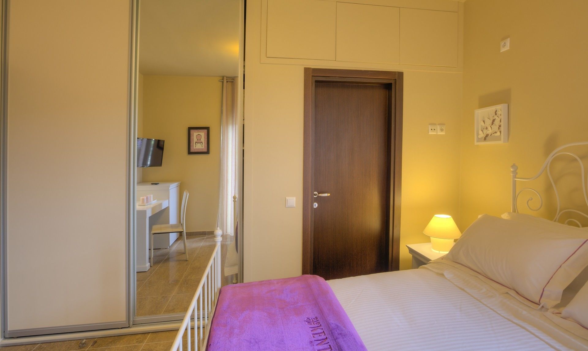 Une chambre d’hôtel avec un lit, une lampe de chevet, une porte en bois, une chaise blanche, une petite table et un téléviseur posé sur un meuble.