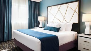 A hotel room with a large bed, two bedside tables with lamps, and a view of the outdoors through a window with blue curtains. - WorldMark Anaheim (Anaheim)