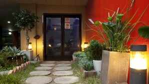 Un chemin menant à l’entrée d’un bâtiment, bordé de plantes en pot et adossé à un mur rouge.