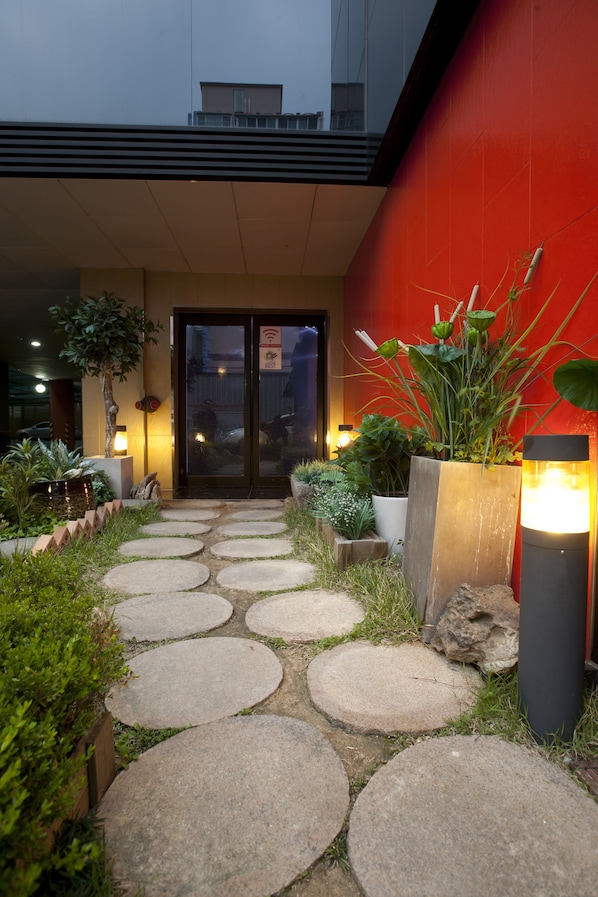 A pathway leading to a building entrance with potted plants and a red wall.