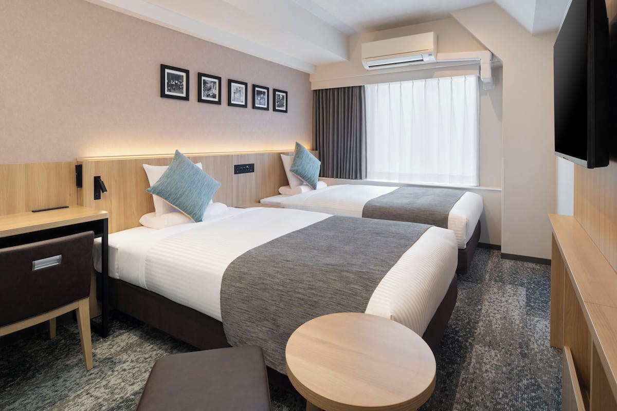 Quarto de hotel com duas camas, uma escrivaninha, uma cadeira, uma mesa redonda pequena e uma televisão.