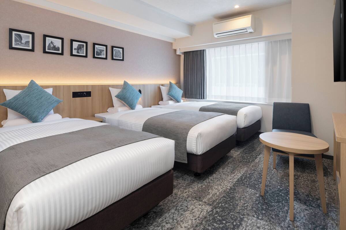 Quarto de hotel com duas camas, uma mesinha, uma cadeira e uma televisão. Há quadros nas paredes e um aparelho de ar condicionado acima da janela.