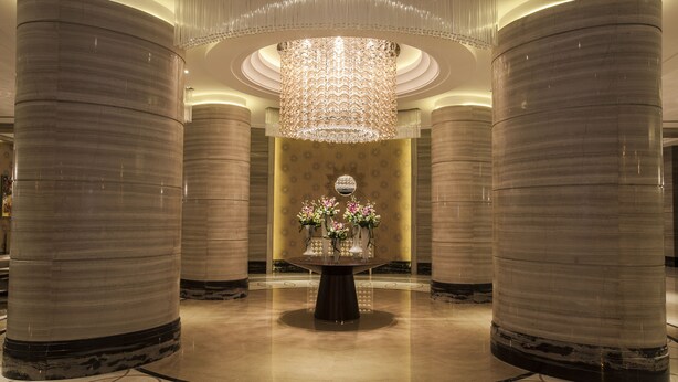 Un lobby espacioso con columnas de mármol, una mesa redonda central con arreglos florales y una lámpara de araña grande y ornamentada.