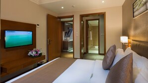 Habitación de hotel con cama, televisor en la pared, escritorio con un jarrón de flores y baño visible a través de una puerta abierta.
