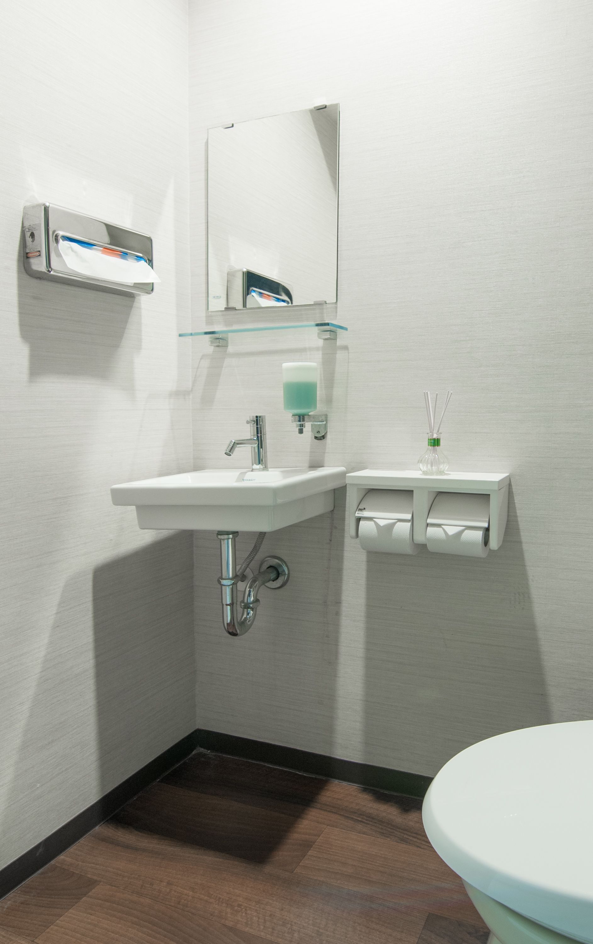 A bathroom with a white sink, mirror, and toilet. There is a soap dispenser and a tissue holder on the wall.