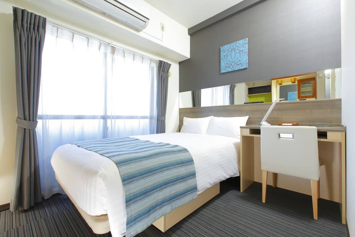 A hotel room with a bed, a desk, and a chair. There is a window with curtains, a wall-mounted air conditioner, and a painting on the wall.