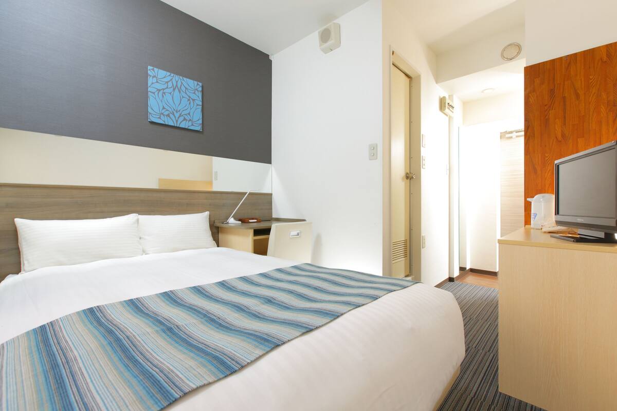 A hotel room with a bed, a desk, a television, and a bathroom visible through an open door.