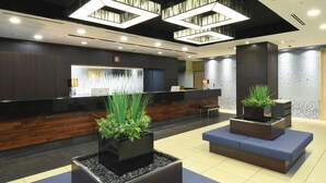 A modern reception area with a wooden reception desk, two blue cushioned seating areas, and decorative planters with green plants. - Hotel MyStays Ochanomizu Conference Center (Tokyo)