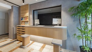 A reception area with a wooden counter, two HP monitors, a plant, and a tiled floor. - Hotel MyStays Nippori (Tokyo)