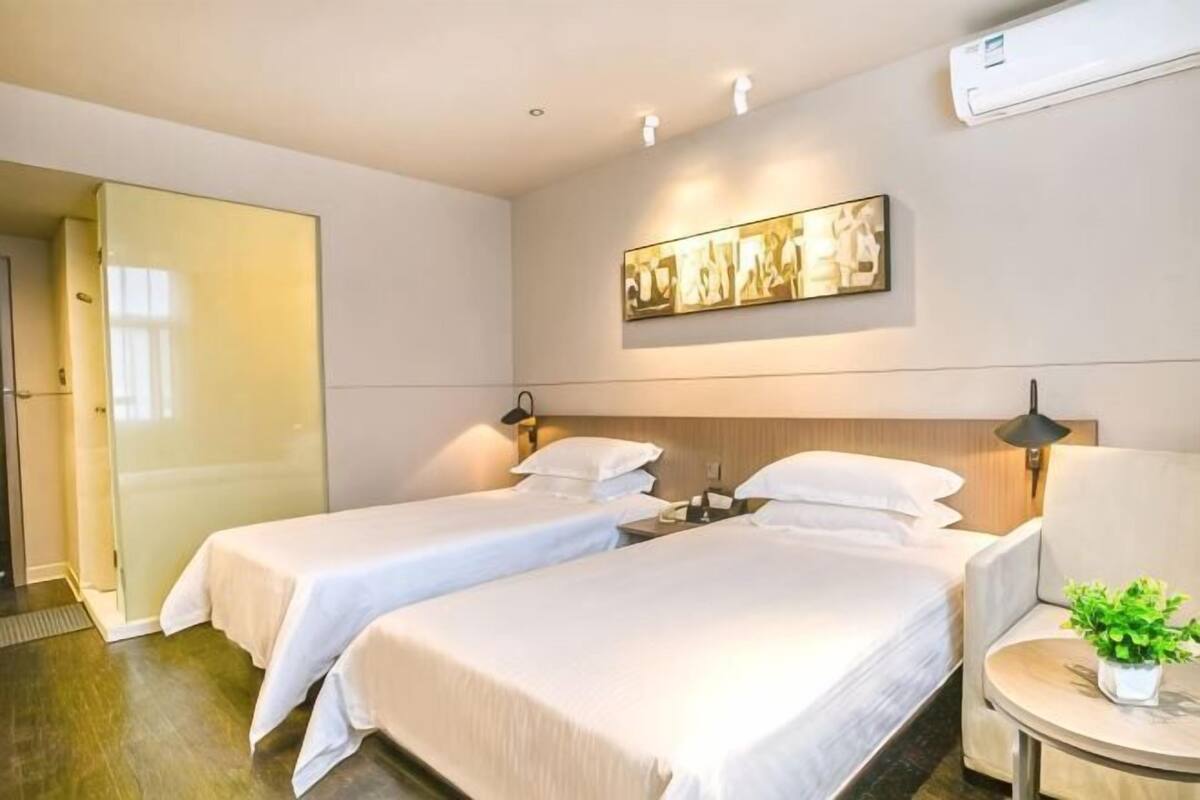 Ein Hotelzimmer mit zwei Betten, einem kleinen Tisch, einem Sessel, einer Topfpflanze und einem Bild an der Wand.