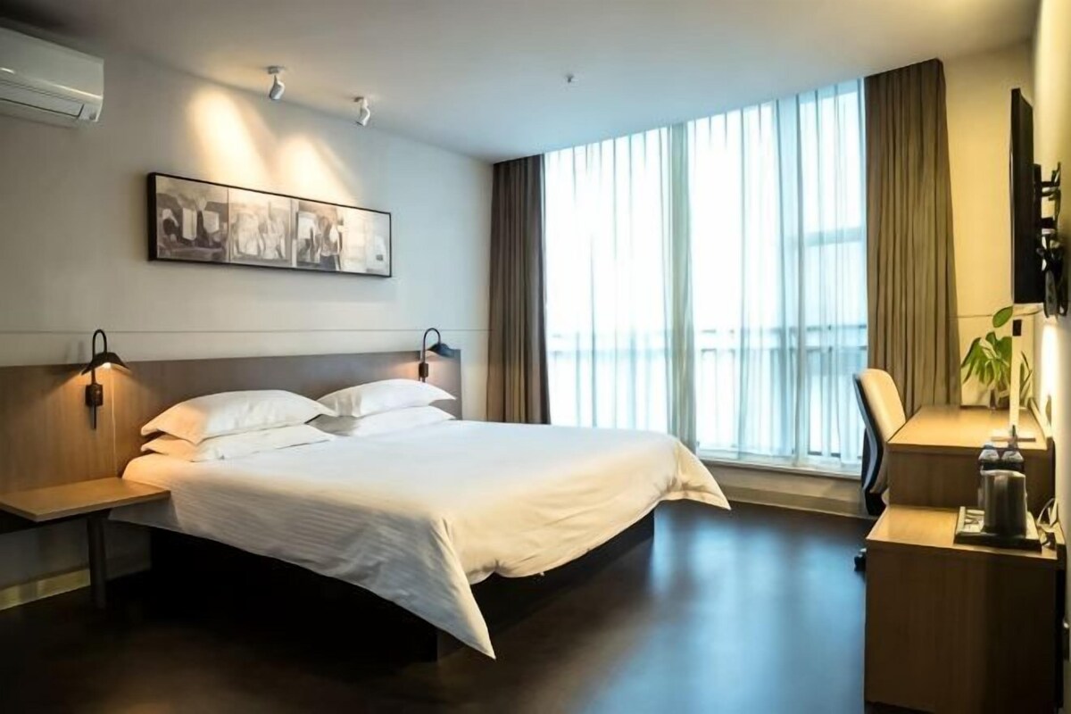 Quarto de hotel com cama, mesas de cabeceira, escrivaninha, cadeira, televisão e uma grande janela com cortinas.
