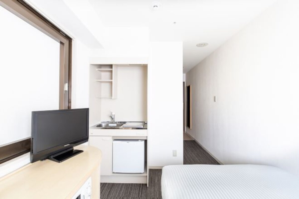 A modern hotel room with a flat-screen TV, a compact kitchenette, a bed, and a large window.