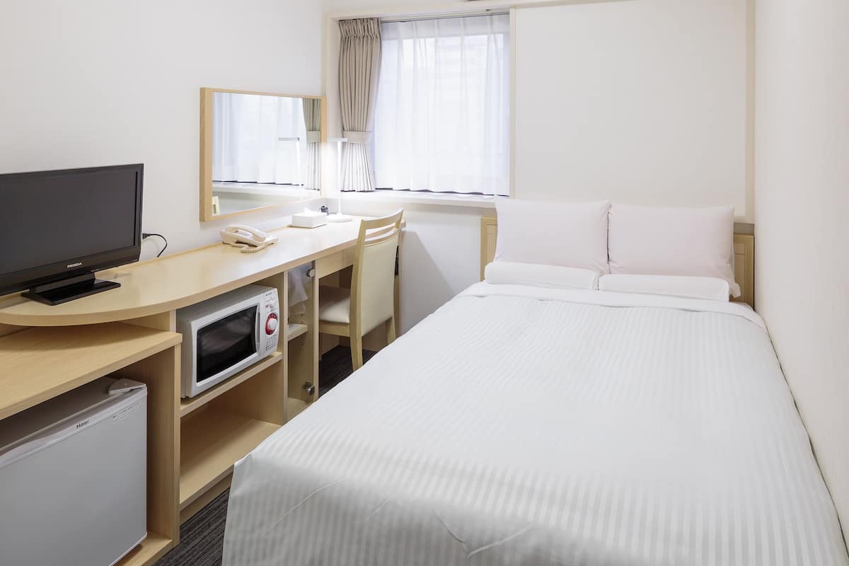 A hotel room with a bed, a desk with a computer and a phone, a television, a microwave, and an air conditioning unit.