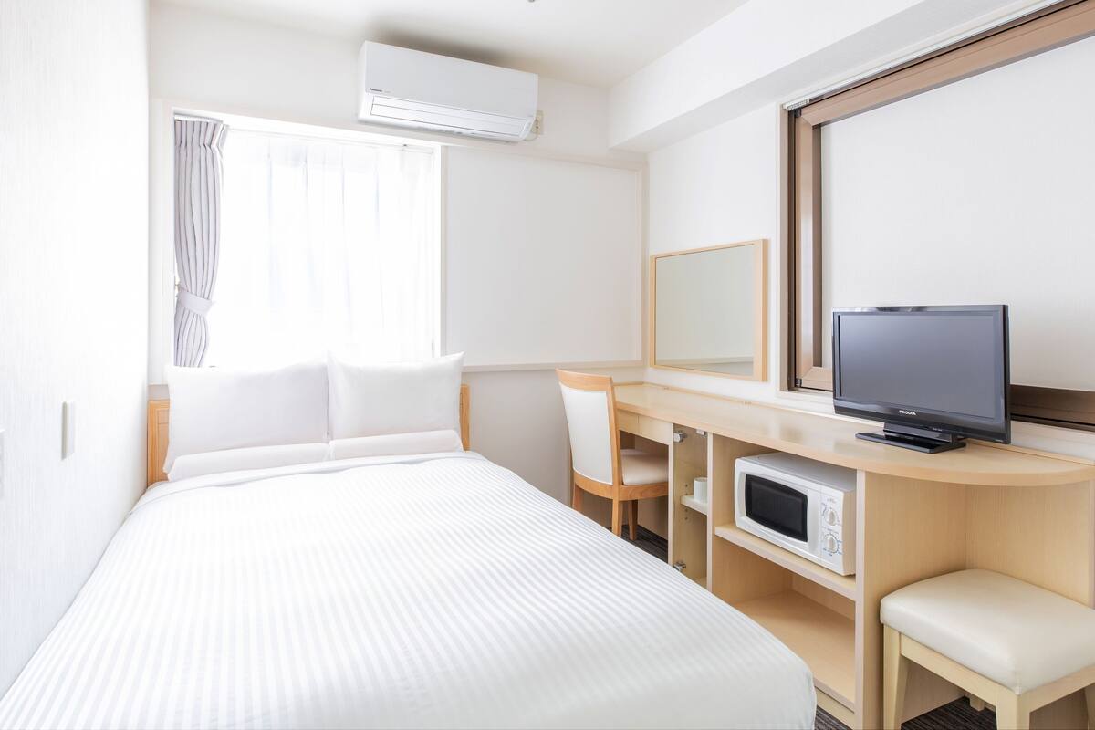A hotel room with a bed, a desk with a TV, a microwave, and an air conditioner.
