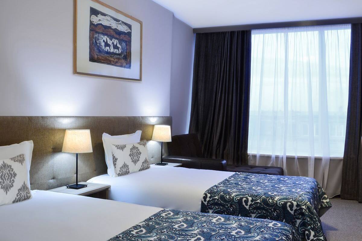 A hotel room with two beds, a headboard, bedside lamps, a large window with curtains, and a framed picture on the wall.